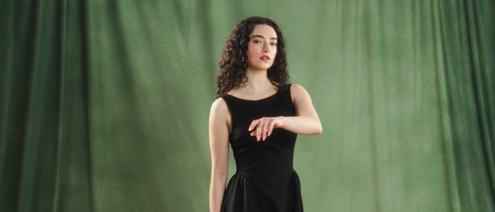 A model in a black dress against a green velvet backdrop, one arm extended