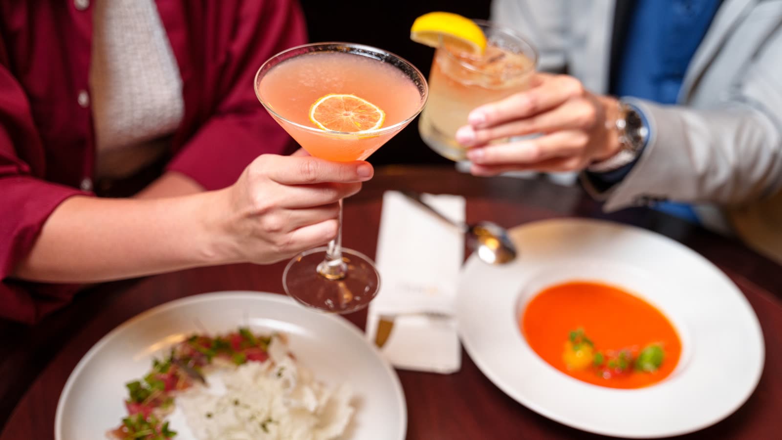 Two diners raise cocktails over plated dishes in a hotel lobby lounge