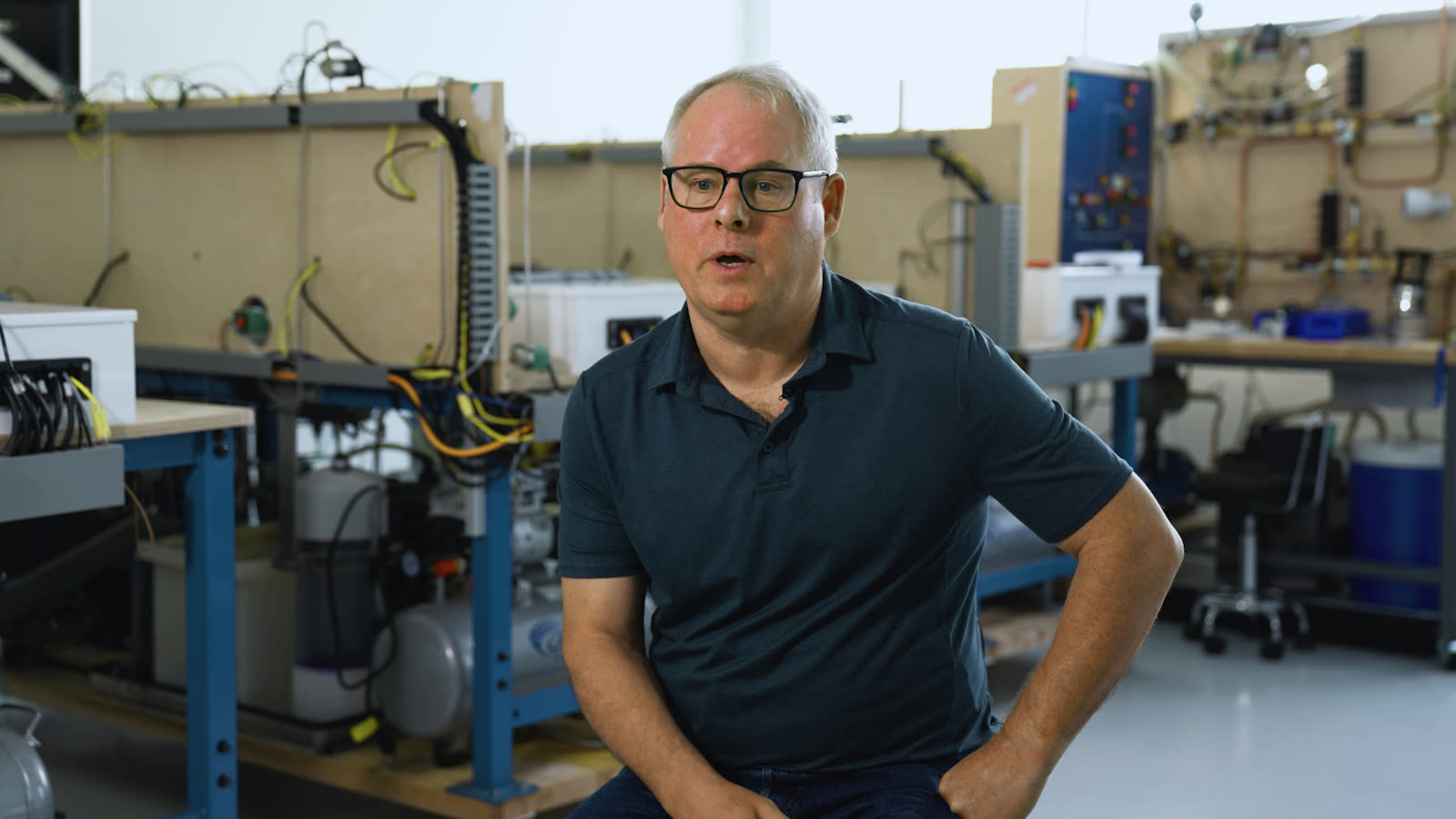 Greg Bohannon speaking, seated with his hands on his knees in front of the lab’s control panels and tubing