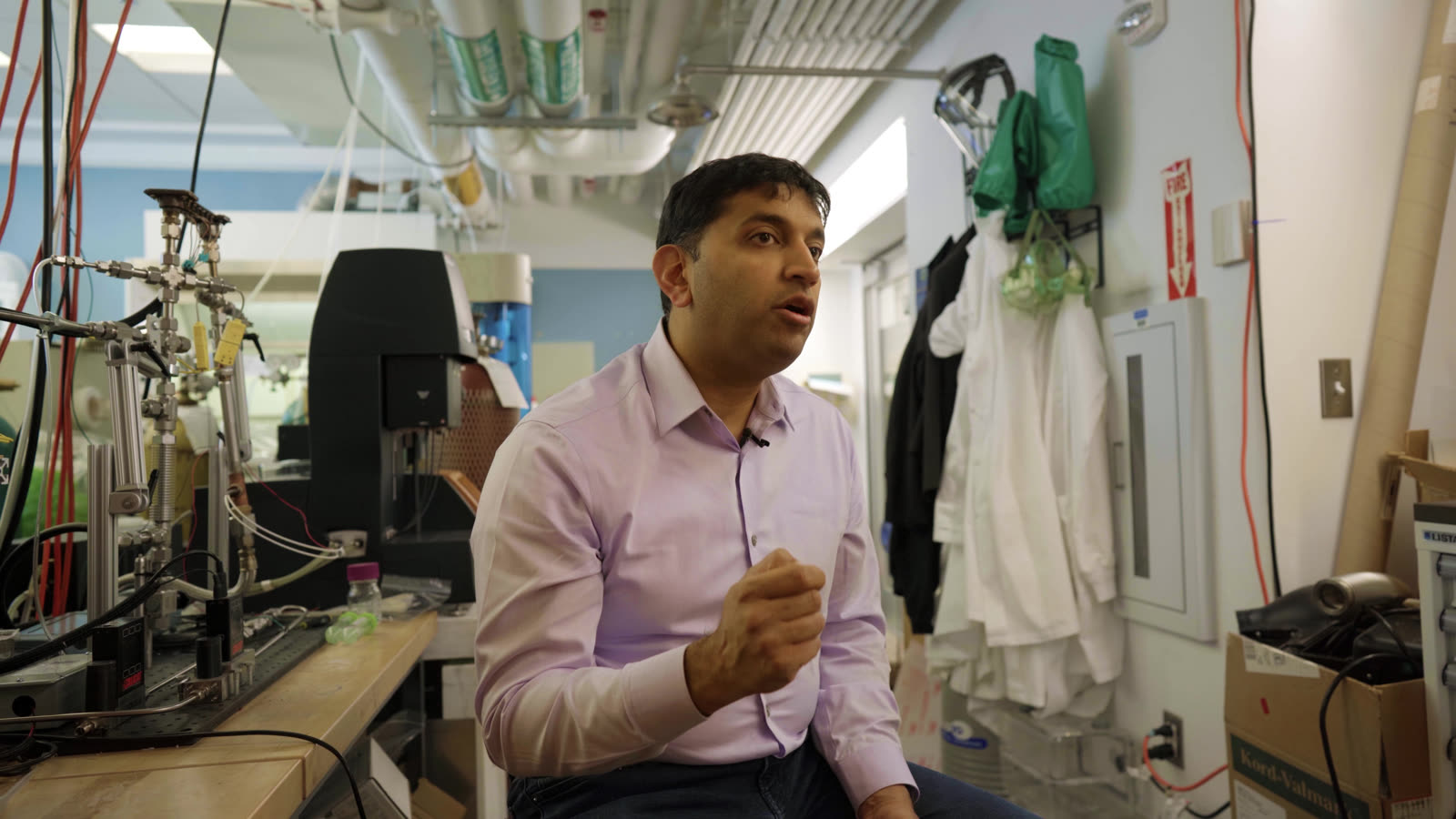 Anurag Bajpayee mid-sentence from the side angle, lab coats and tubing behind him