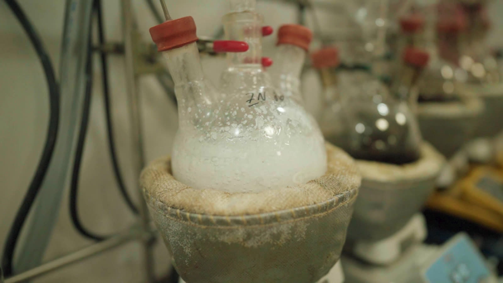 Close-up of a multi-neck reaction flask seated in a heating mantle, the liquid inside pale and clouded