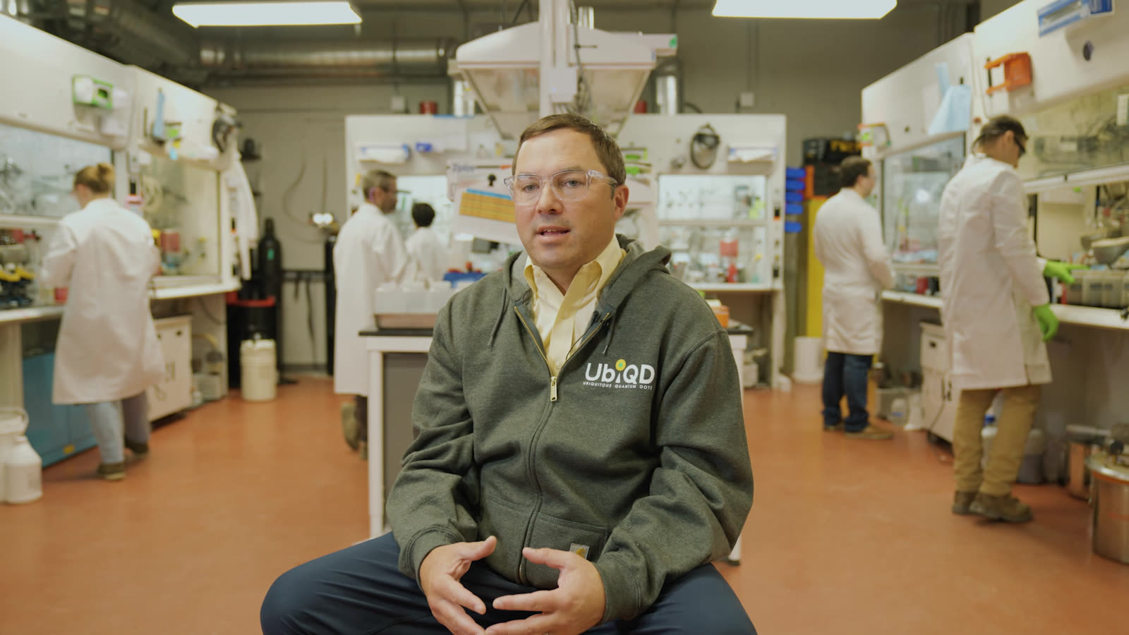 Hunter McDaniel speaking, seated in the centre aisle of the lab with white-coated scientists working at the benches behind him